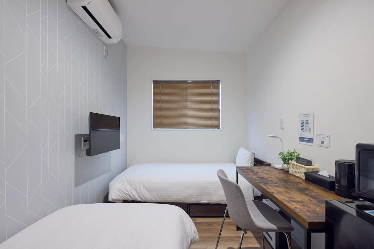 A small, modern room with two beds, a wooden desk, a television, and a window with blinds.