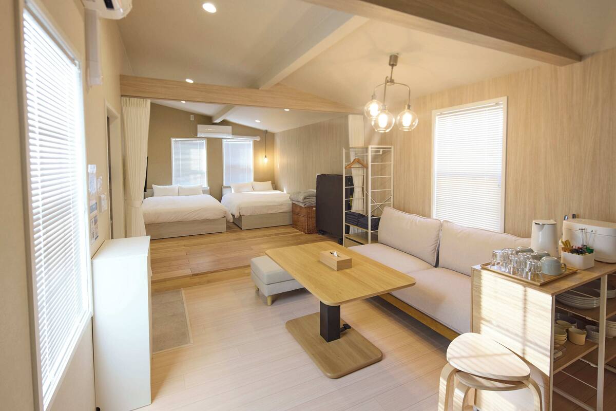 A modern hotel room with a sofa, a small dining table, a kitchenette, and a separate bedroom with a bed.