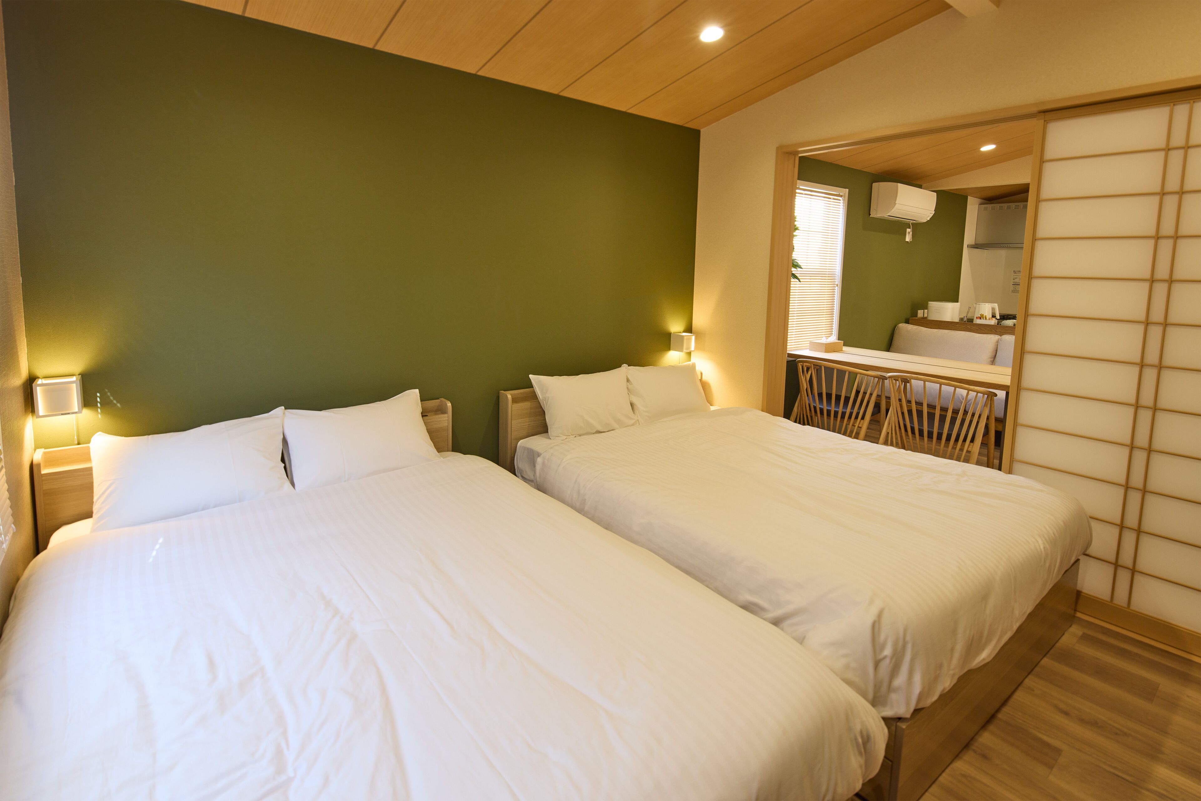 A hotel room with two beds, a wooden ceiling, a green wall, and a mirror reflecting a bathroom.