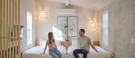 Un dormitorio con dos camas individuales, un separador de listones de madera, un aire acondicionado instalado en la pared y una obra de arte enmarcada sobre las camas.