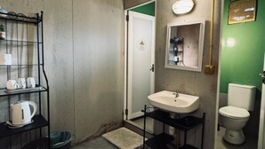 A compact bathroom with a white sink, a mirror, and a toilet. There is a black shelf with cups and a kettle, and a green trash bin. - Padi Box (Sekinchan)