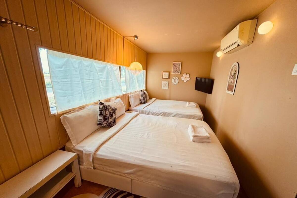 A bedroom with two beds, a bench, a wall-mounted TV, and a window with a curtain.