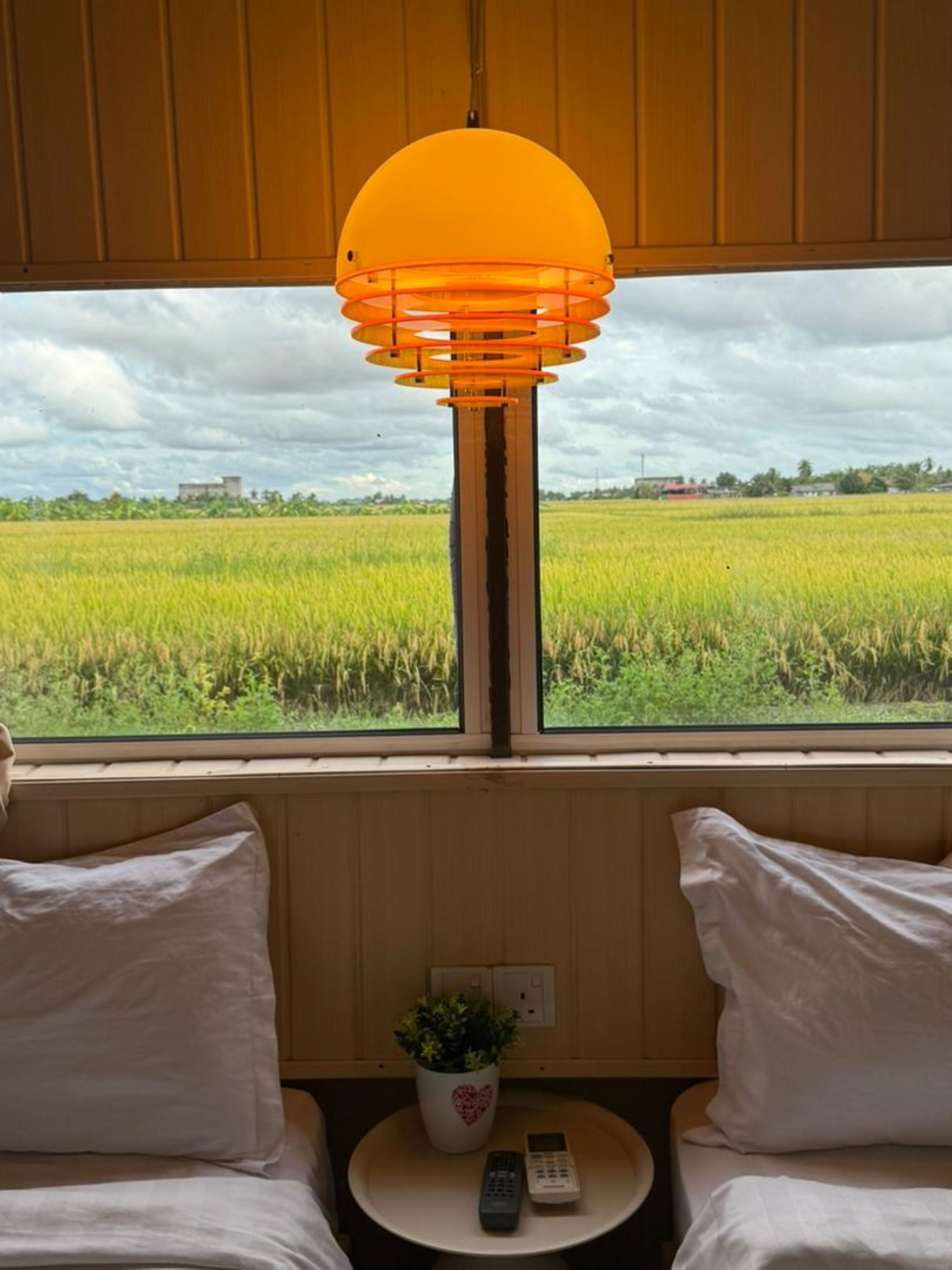A room with a view of a green field through a window, a round table with a remote control and a potted plant, and a bed with white bedding.