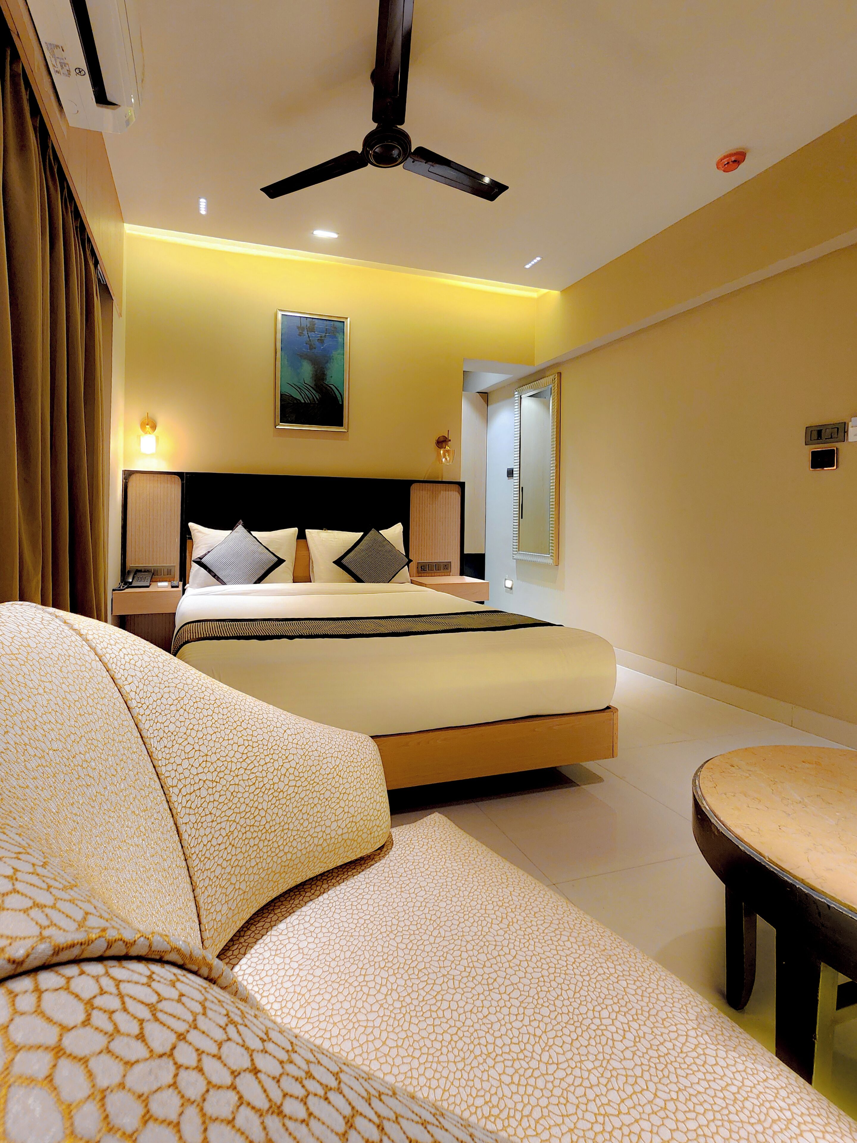 A hotel room with a bed, a ceiling fan, a bedside table with a lamp, and a view of the outside.