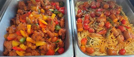 Two metal food dispensers with stir-fried meat and vegetables, and spaghetti with meatballs and cherry tomatoes.