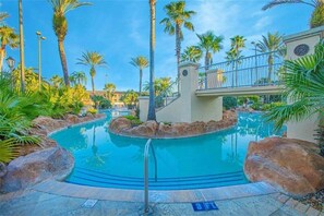 A swimming pool surrounded by palm trees and rocks, with a bridge and a clock tower in the background. - 1307-4-ca-townhome at Regal Palms (Davenport)