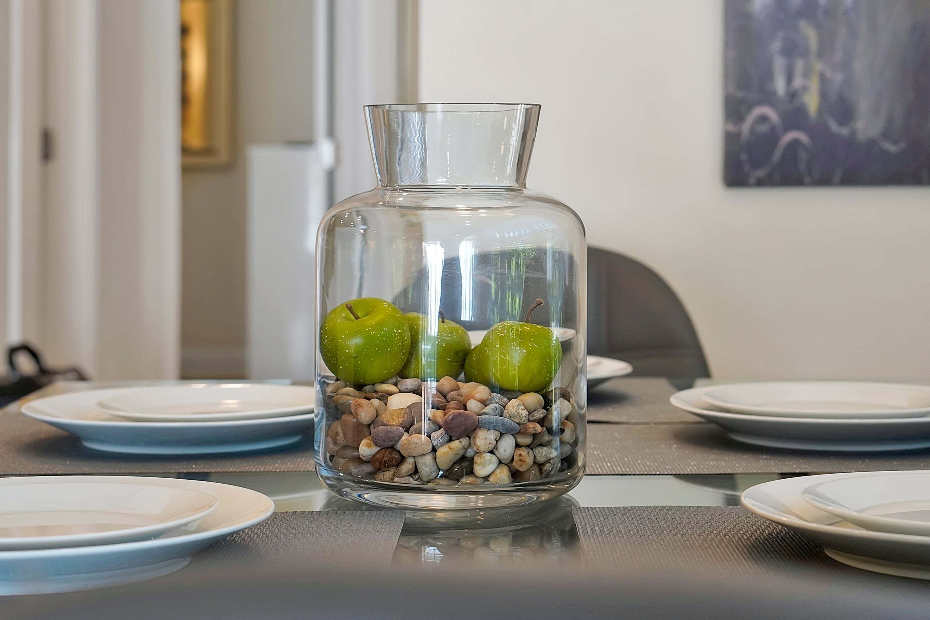 A clear glass vase with green apples and pebbles on a table.