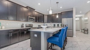 Una cocina moderna con armarios oscuros, electrodomésticos de acero inoxidable, una isla central con taburetes y luminarias colgantes.
