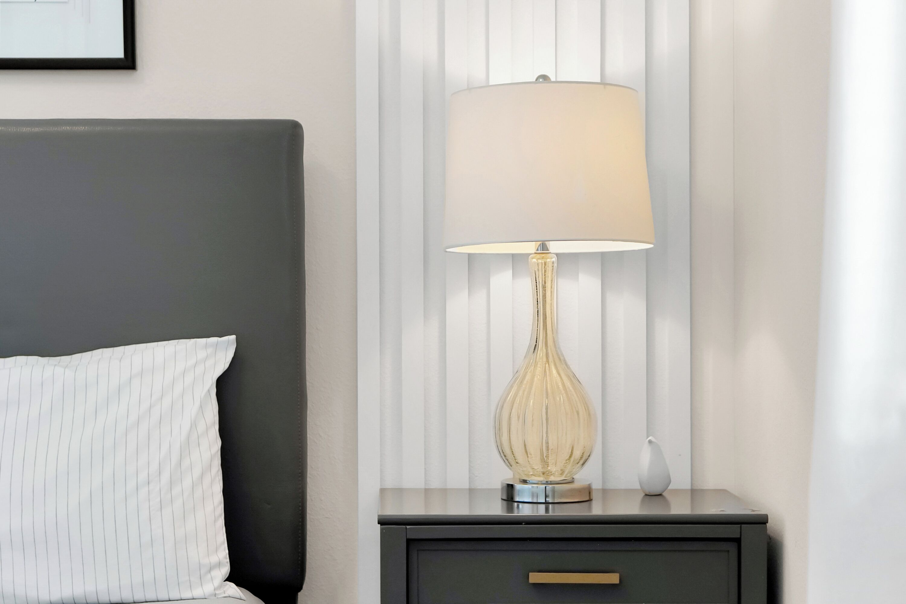 A neatly made bed with a white pillow and a dark headboard. A bedside table with a lamp and a small vase is visible.