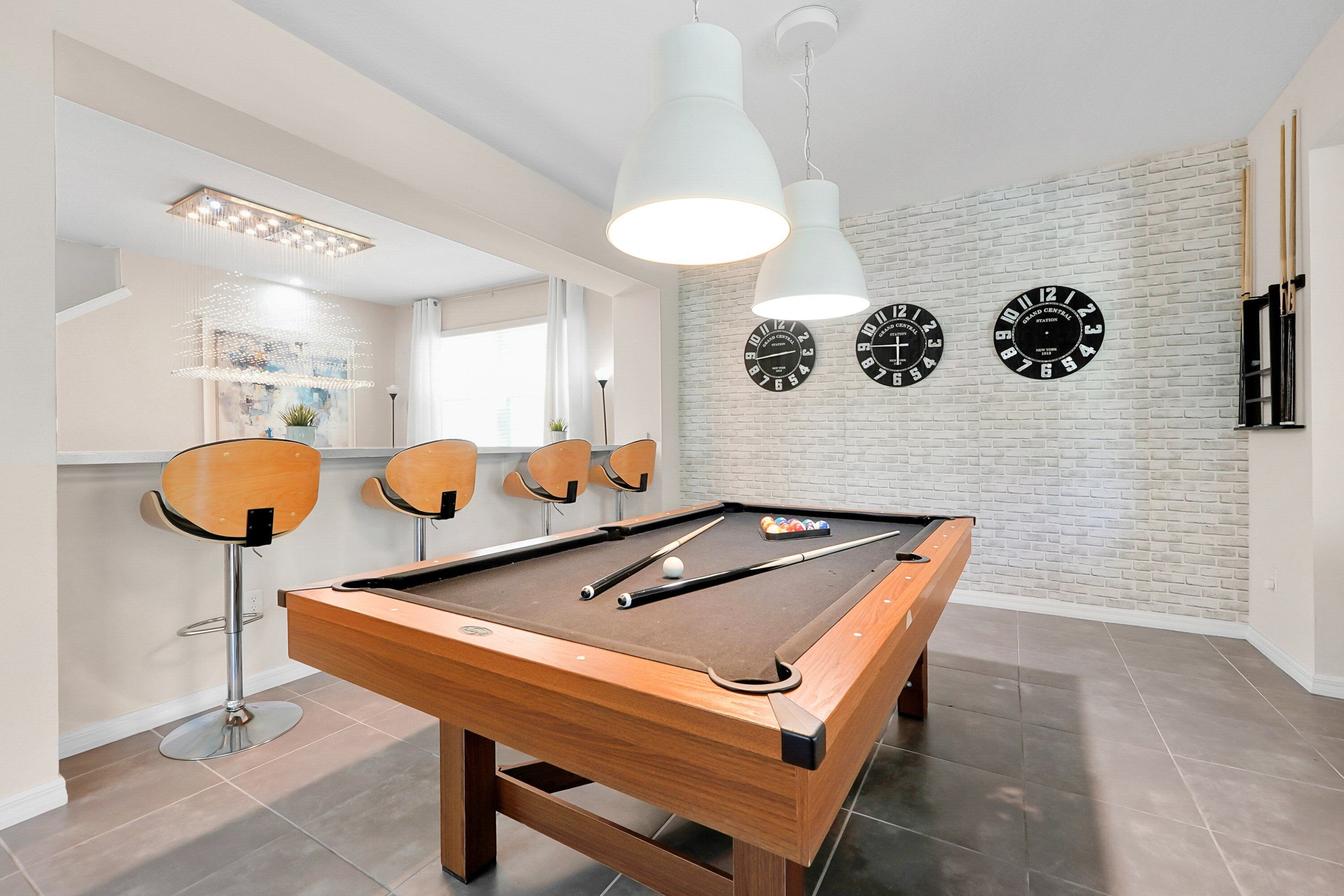 A pool table with four chairs, a billiards cue, and three clocks on the wall.