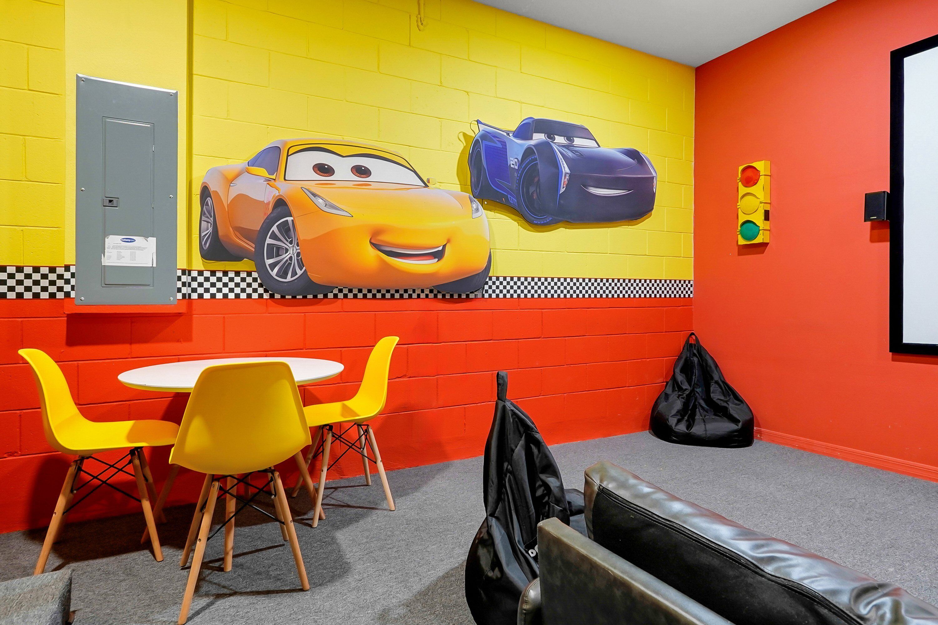 Um quarto com parede de tijolos amarelos, um mural de carros animados, uma mesa branca com cadeiras amarelas e uma bolsa preta no chão.