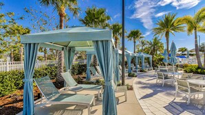 Een overdekt terras met lounge stoelen en parasols, omgeven door palmbomen en een witte hek.