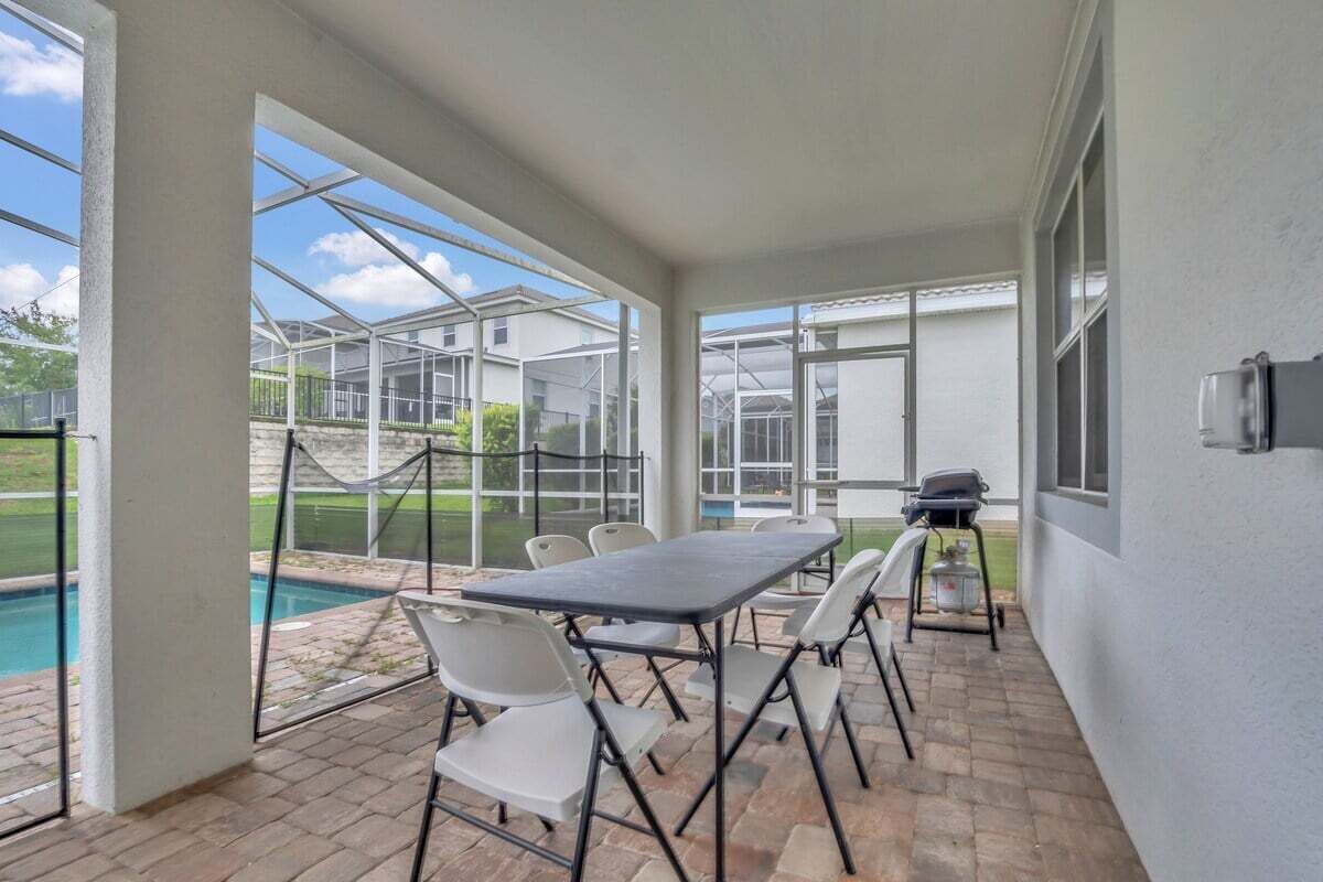 A covered patio area with a table and chairs, a grill, and a view of a swimming pool and greenhouse structures.