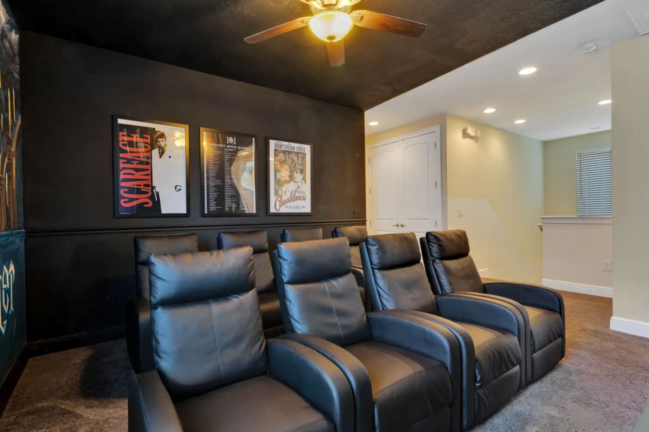 A home theater room with black leather reclining chairs, a ceiling fan, and movie posters on the wall.