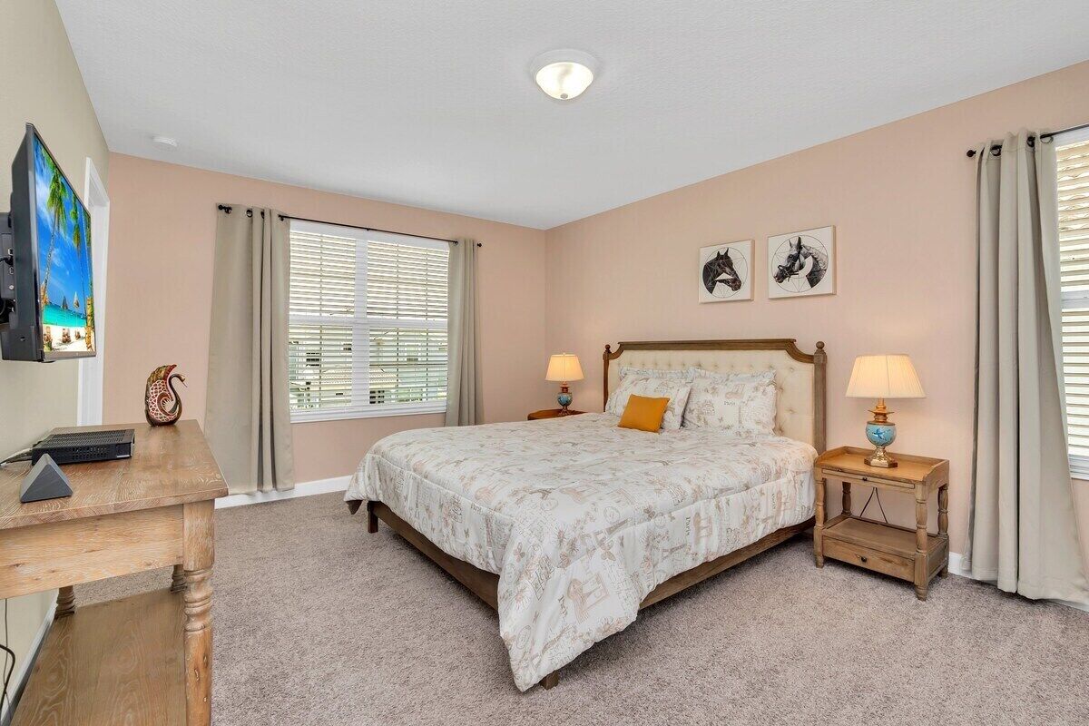 A bedroom with a bed, two bedside tables with lamps, a desk with a computer, a TV, and a window with blinds.