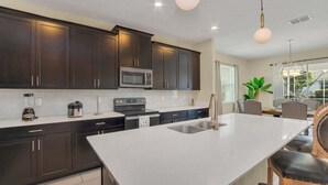 Una cocina moderna con armarios oscuros, una isla grande con pileta, microondas y estufa. Se ven sillas y una mesa de comedor al fondo.