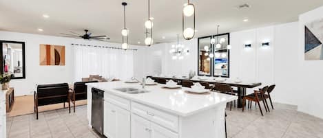 Una cocina moderna con armarios blancos, una isla central, iluminación colgante y un comedor con sillas y una mesa.