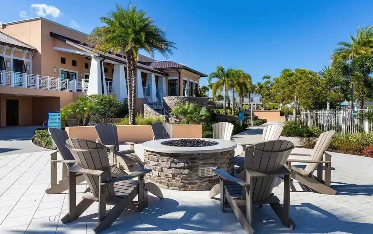 An outdoor seating area with Adirondack chairs and a stone fire pit, surrounded by palm trees and a building with a balcony.