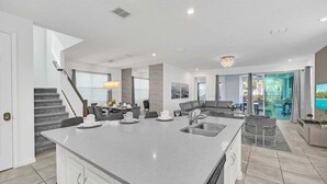 A modern kitchen with a central island, a dining area with a sofa and table, and a staircase leading to another level. - 8939ad · Amazing Solara 7br/ Pool /spa /free Waterpark (Kissimmee)