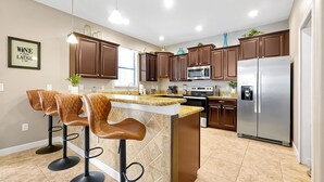 A modern kitchen with brown cabinets, a stainless steel refrigerator, a microwave, and a bar area with two stools. - 1452 Myrtlewood, 6 Beds (Davenport)