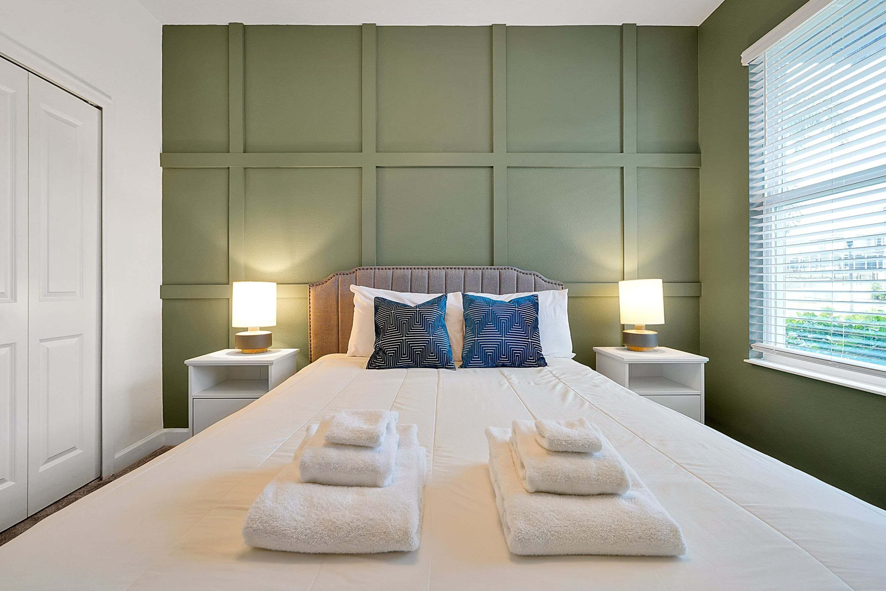 A hotel room with a large bed, two bedside lamps, a headboard, and a window with blinds.