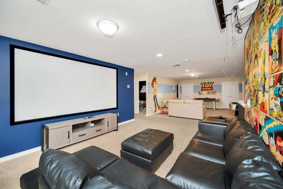 A living room with a large TV, black leather sofas, a kitchen in the background, and comic book-themed wall art.