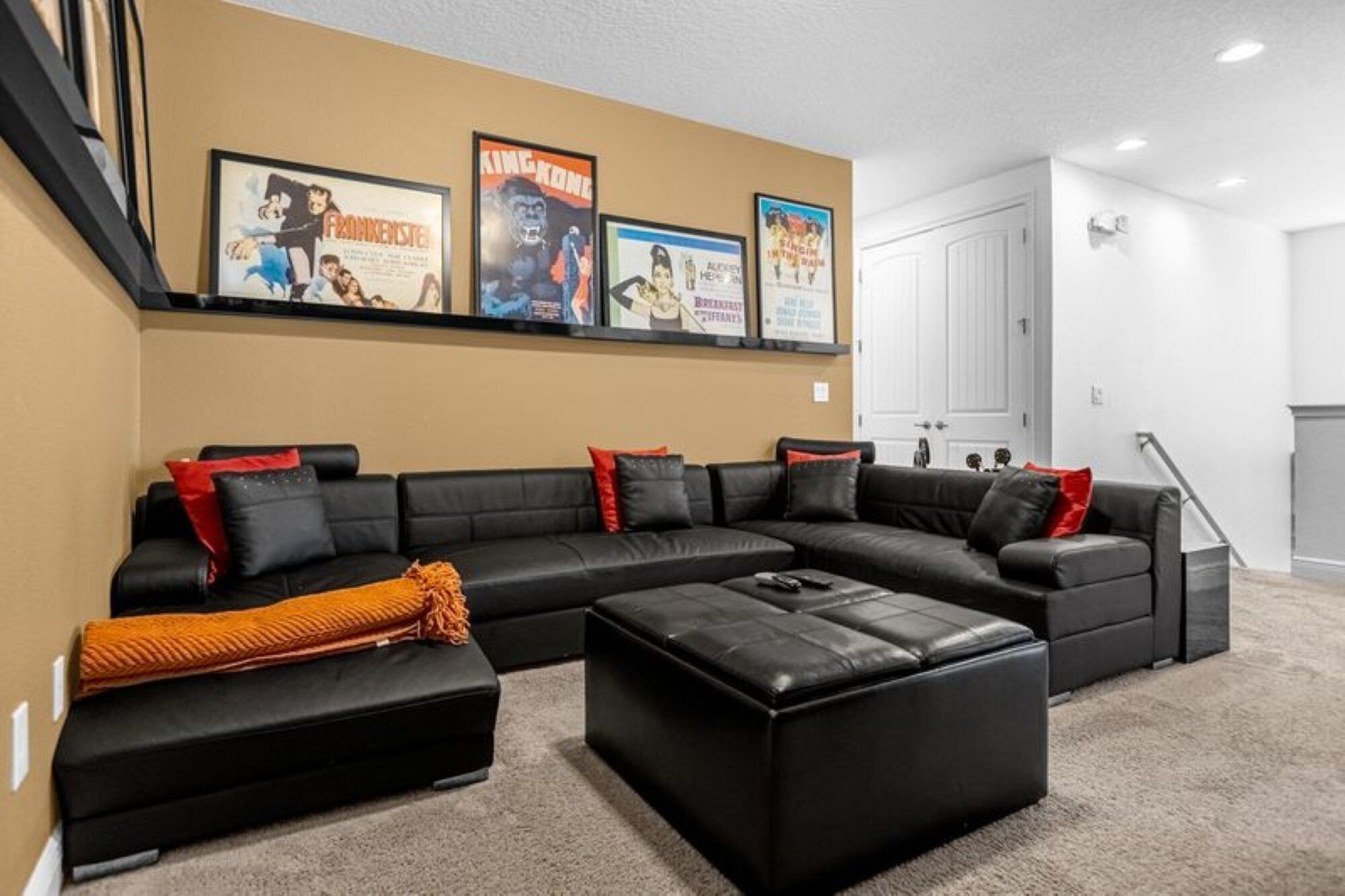 A living room with a black sectional sofa, a black ottoman, and a red throw blanket. There are framed movie posters on the wall.