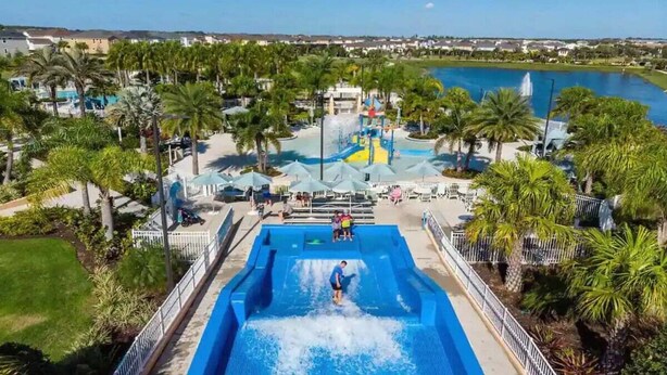 A water park with a large pool, palm trees, and a view of a lake and buildings in the background. - 8950cbd · Amazing 9BR Lakeview Villa in Solara With Pool/spa (Kissimmee)