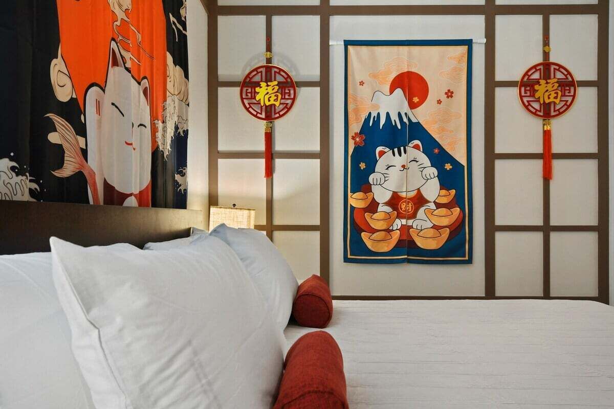A hotel room with a bed, pillows, a wall with traditional Japanese artwork, and two red hanging decorations.