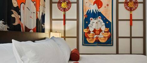 A hotel room with a bed, pillows, a wall with traditional Japanese artwork, and two red hanging decorations.
