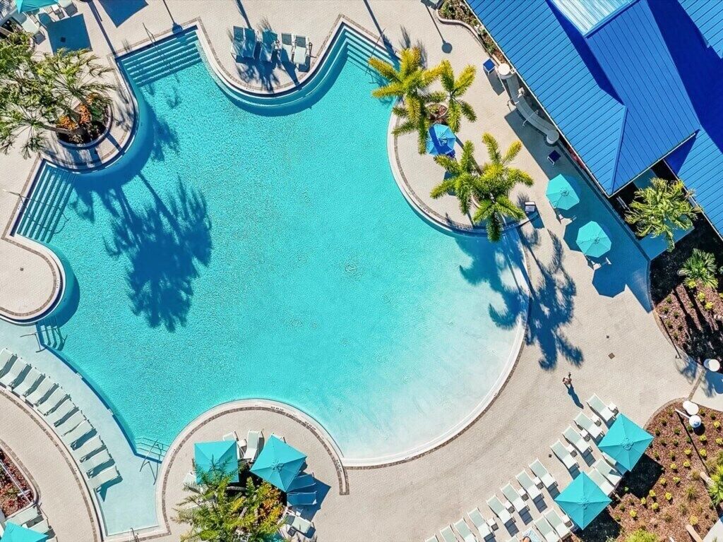 An aerial view of a swimming pool with sun loungers and umbrellas, surrounded by palm trees and a building with a blue roof.