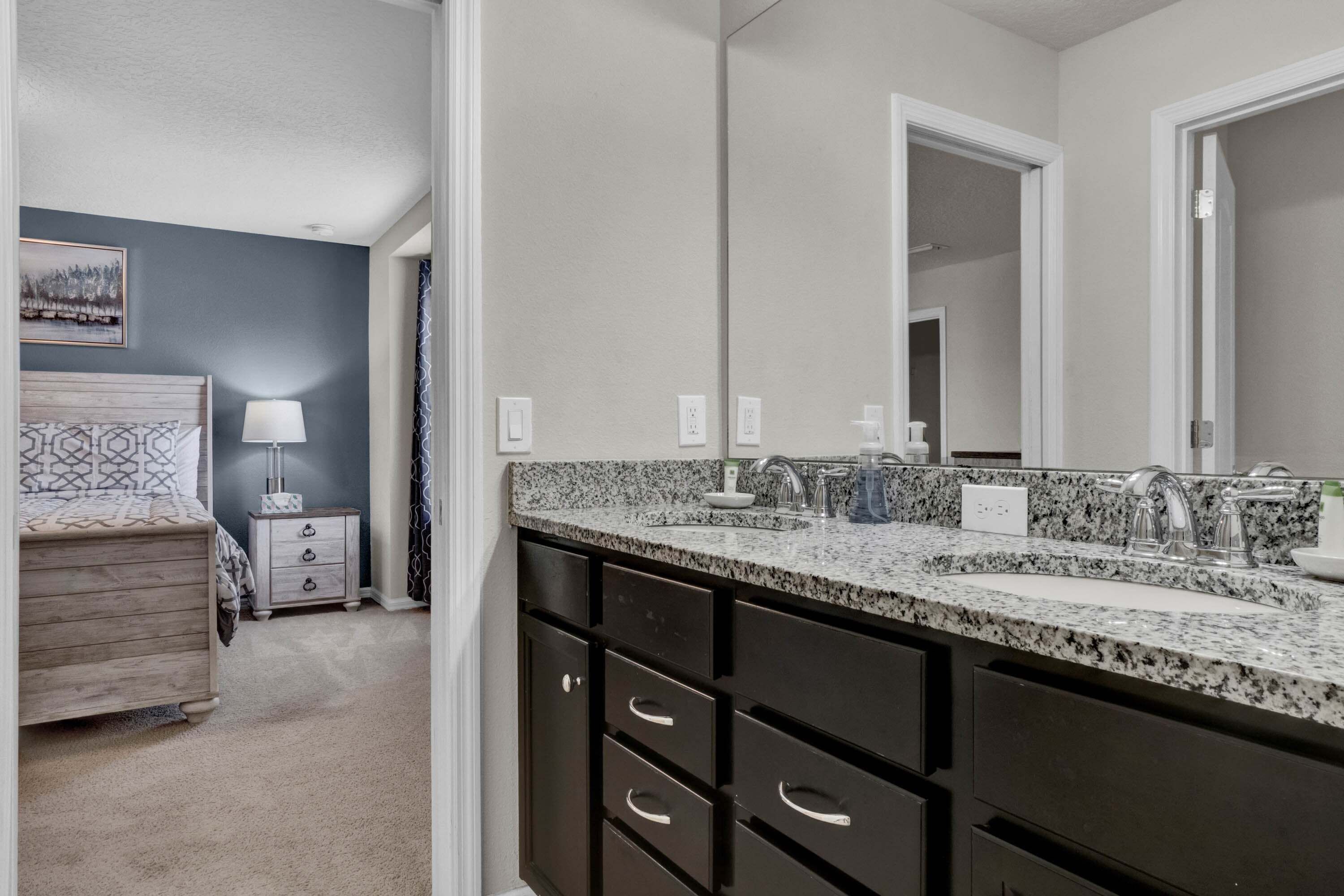 Une salle de bain avec un comptoir en granit, des meubles sombres, un grand miroir, et une chambre adjacente avec un lit doté d’une tête de lit à motifs.
