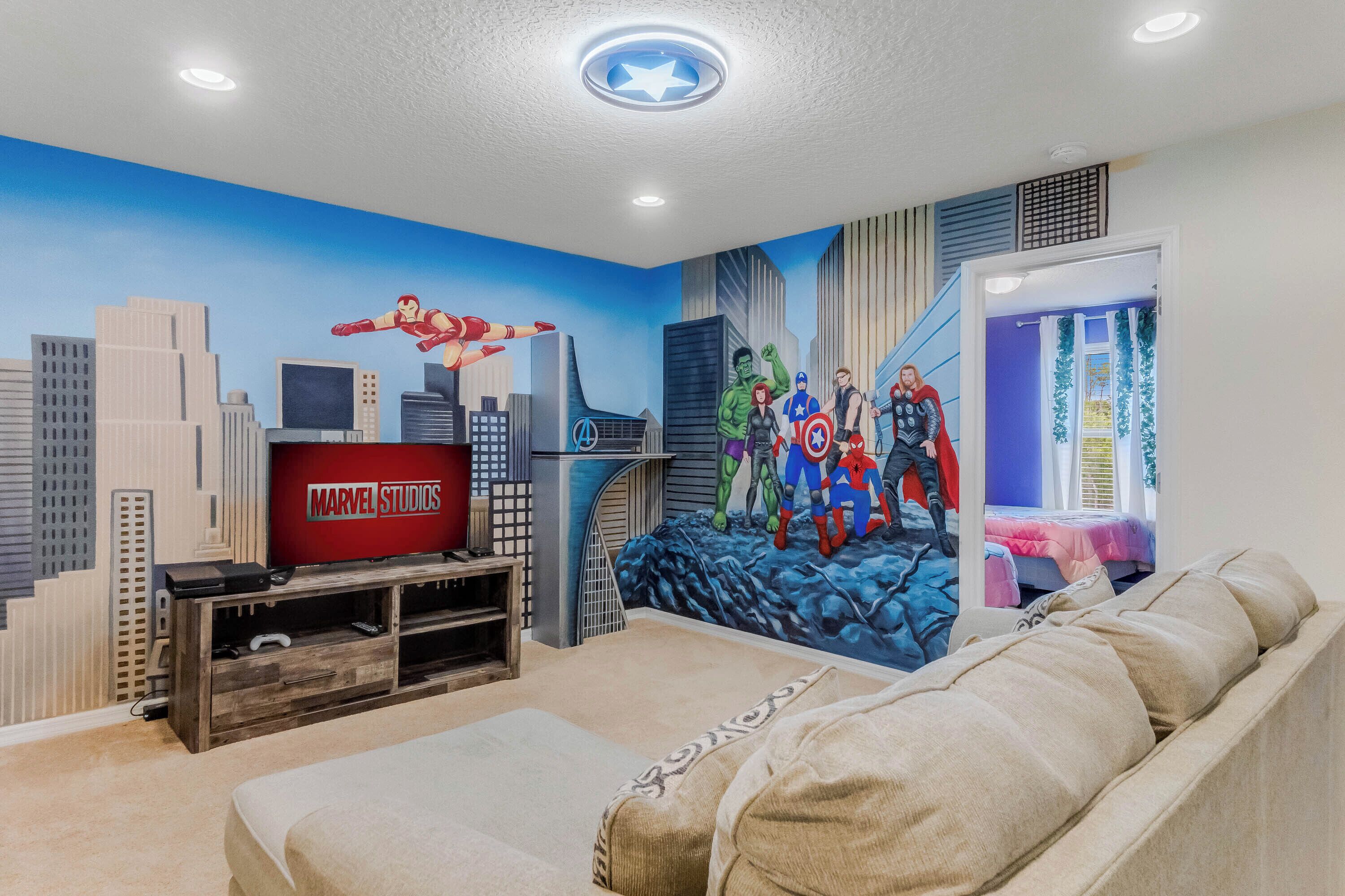 A room with a large wall mural of Marvel characters, a television, and a bed.