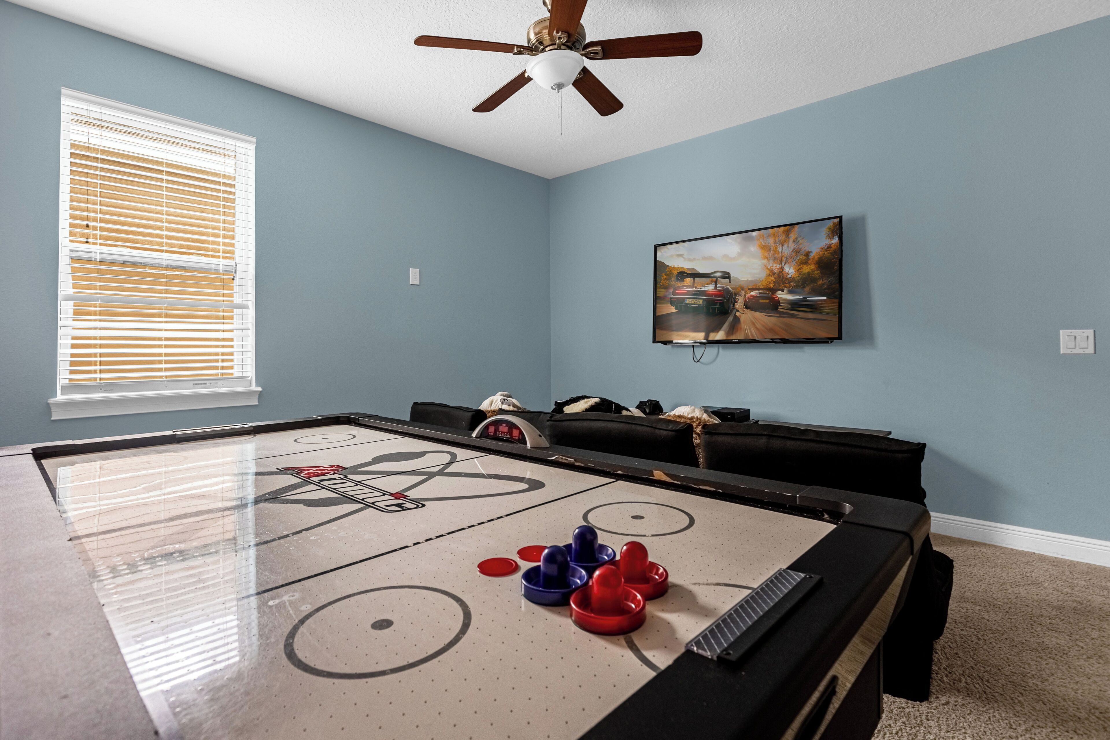 Una habitación con mesa de juego de pachín, ventilador de techo, ventana con persianas y un cuadro en la pared.