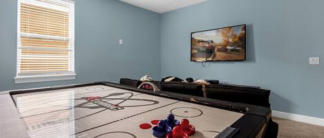 Una habitación con mesa de juego de pachín, ventilador de techo, ventana con persianas y un cuadro en la pared.