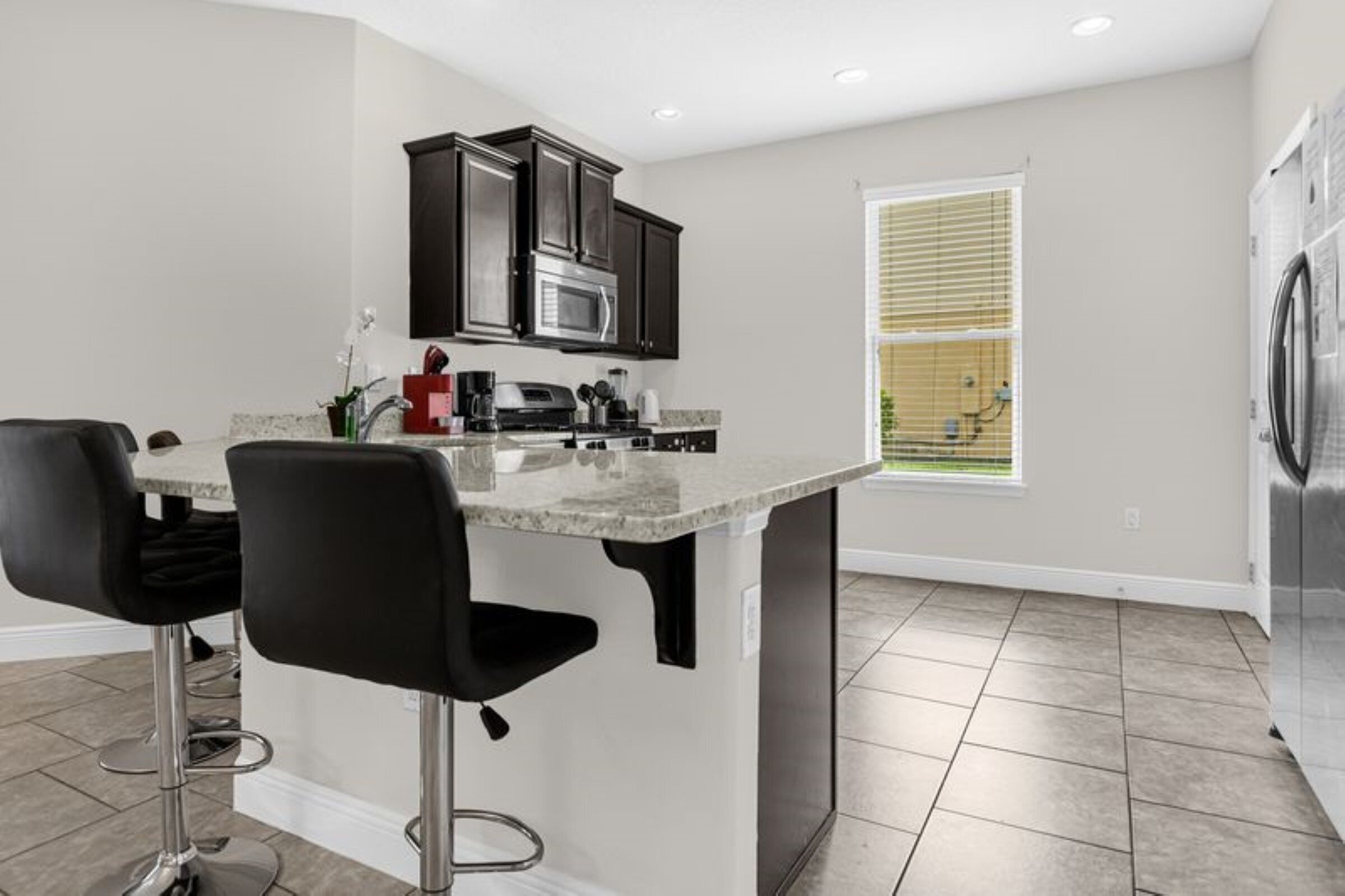 A modern kitchen with dark cabinets, a central island with bar stools, a built-in microwave, and a large window with blinds.