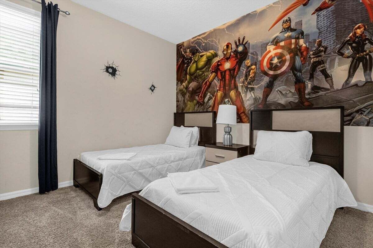 A hotel room with two beds, a large superhero-themed mural on the wall, a window with curtains, and a bedside table with a lamp.