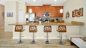A modern kitchen with wooden cabinets, stainless steel appliances, and a granite countertop. There are three bar stools with a unique design. - 1576 Moon,champions Gate,4bed (Davenport)