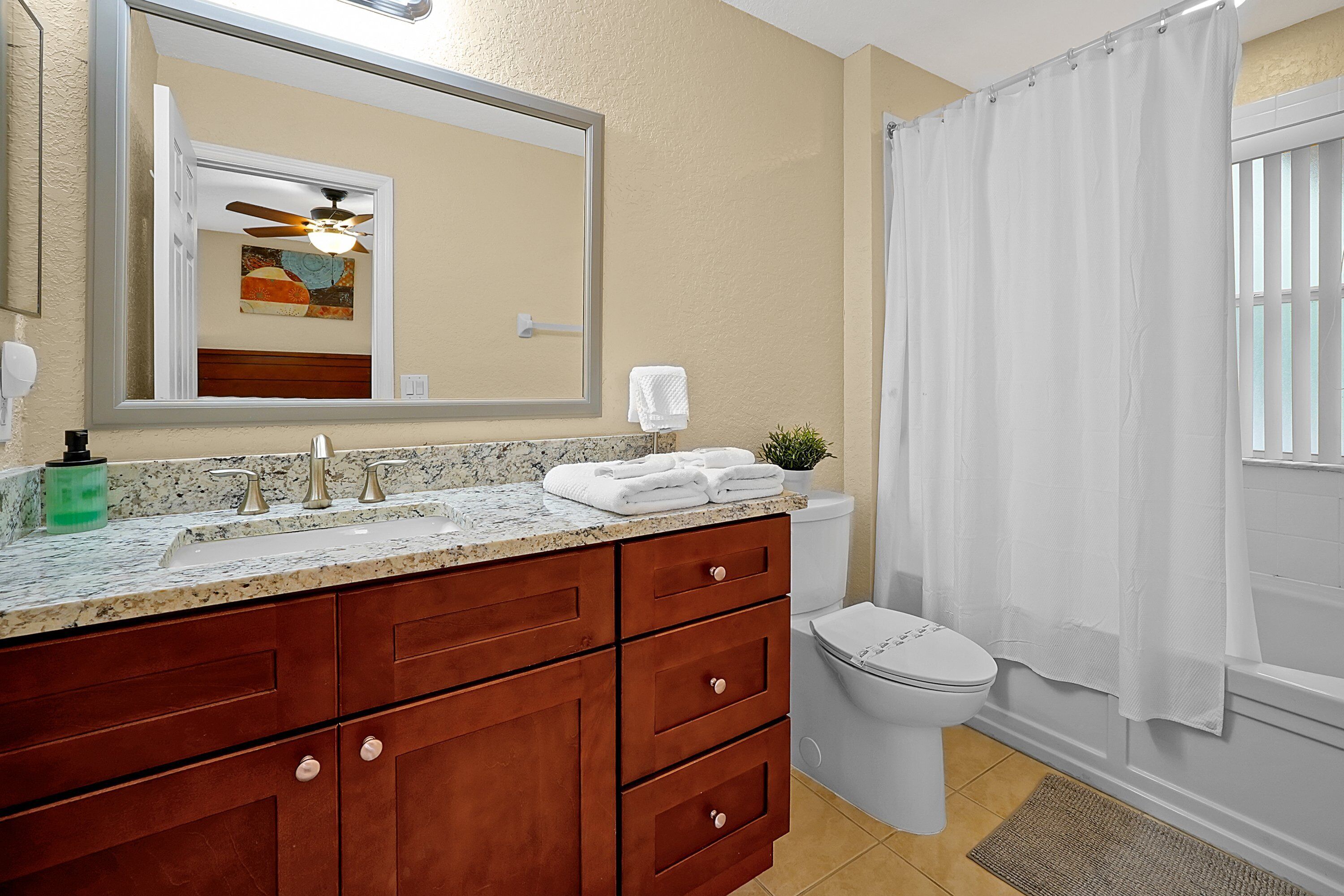 Une salle de bain avec un lavabo en marbre, un meuble-lavabo en bois, un ventilateur de plafond, des toilettes et un rideau de douche.
