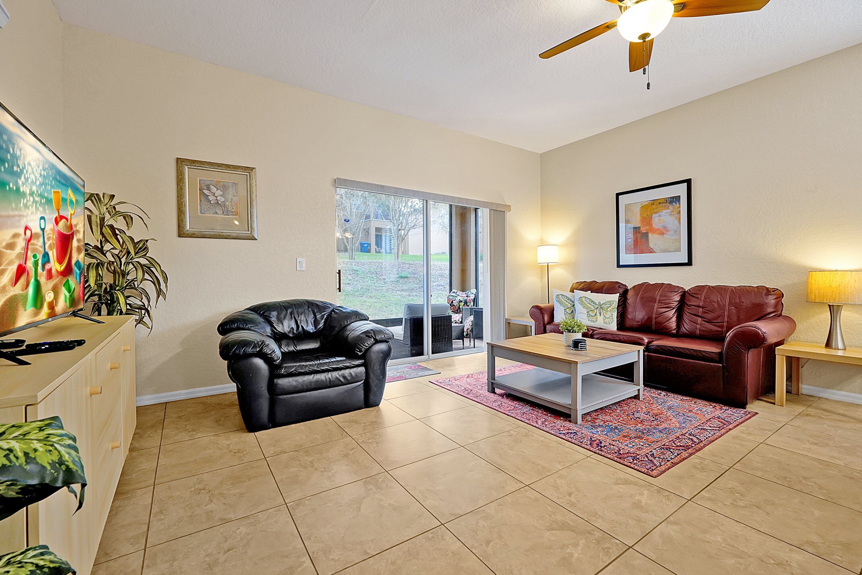 A living room with a black leather chair, a brown leather sofa, a coffee table, and a TV. There is a ceiling fan, a window with a view, and a door leading outside.