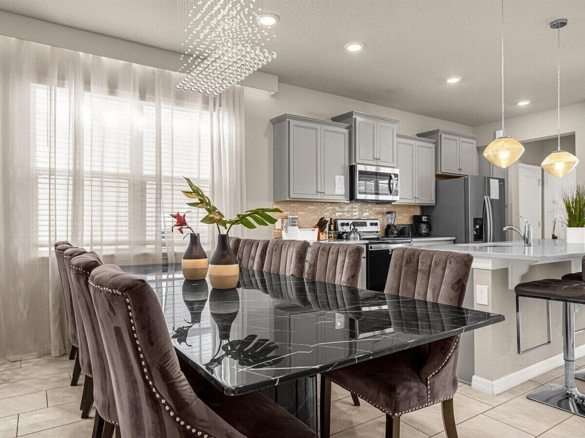 A modern kitchen with grey cabinets, a black marble island, and a dining area with a glass table and cushioned chairs.