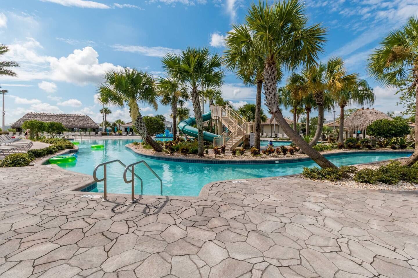A resort pool area with a central water slide, palm trees, and a thatched-roof building.