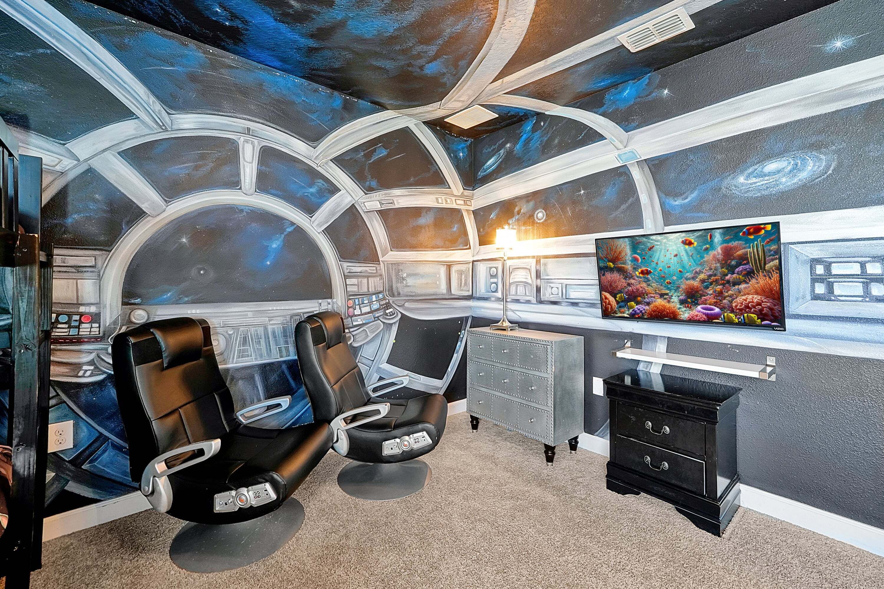 A room with a space-themed mural, two reclining chairs, a desk with a computer, and a television.