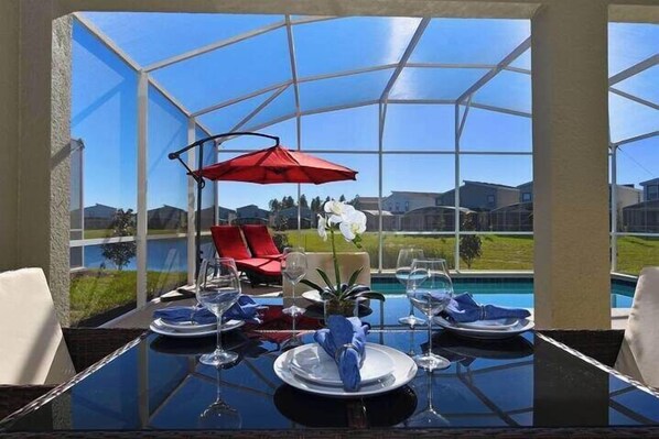 A dining area with a glass table, red umbrella, and a view of a swimming pool and houses. - 8972ccd · Amazing 6Br Villa Near Disney With Lake View (Kissimmee)