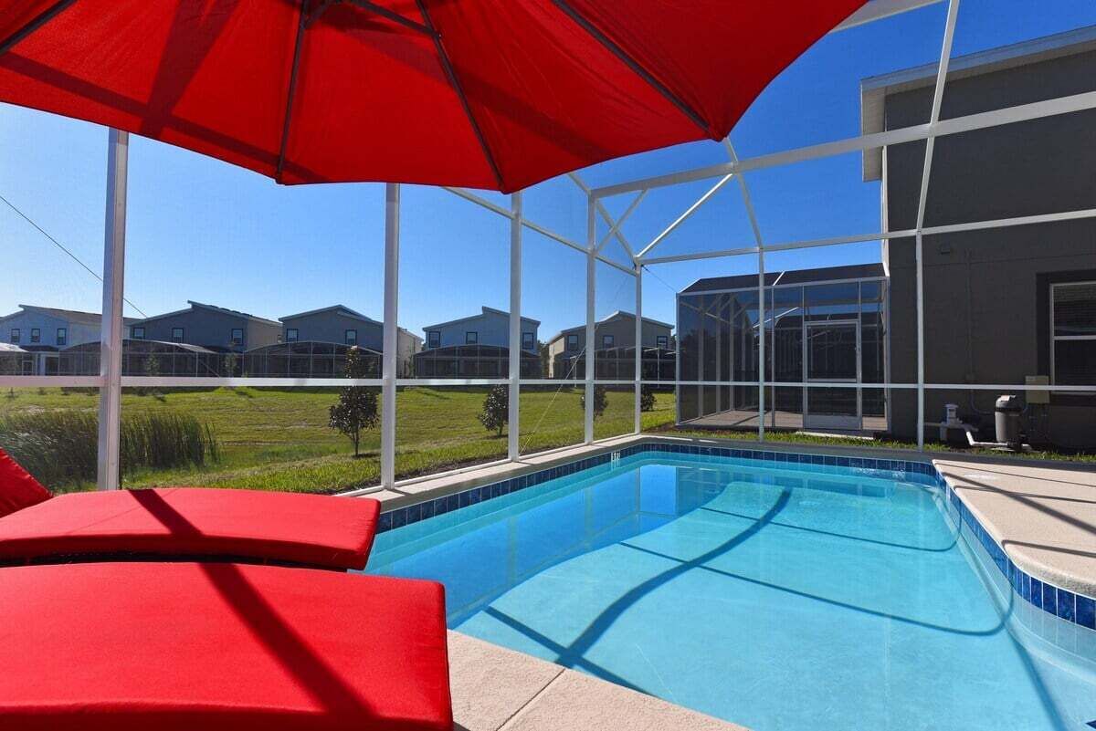 Piscina con sillas y una sombrilla rojas, rodeada por un recinto de cristal con vistas a casas.