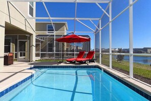 A swimming pool with red lounge chairs and an umbrella, surrounded by a glass enclosure with a view of a lake and buildings. - 8972ccd · Amazing 6Br Villa Near Disney With Lake View (Kissimmee)