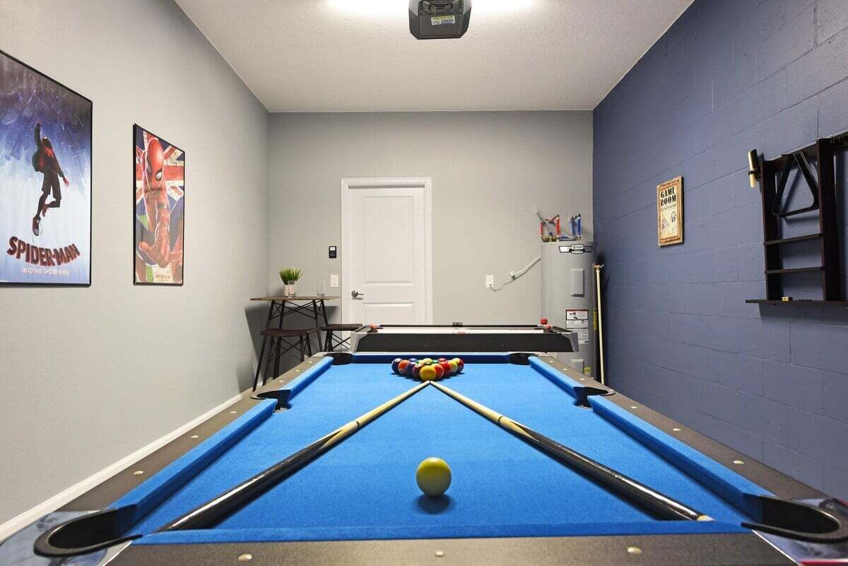 Een biljart met een wit speelbal, twee biljartkeus en een set ballen. Er is een deur, een klein tafeltje met een plant en een poster aan de muur.