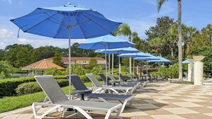 A row of sun loungers with blue umbrellas, arranged on a checkered tile floor, with a palm tree and a building in the background. - 8567 BAY Lily Loop, 3 BED (Kissimmee)