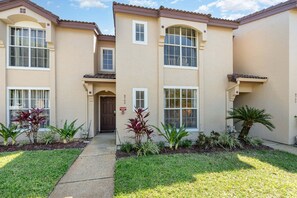 A two-story residential building with a beige exterior, numbered 872, featuring a front door, windows, and a small garden with plants and a palm tree. - 8702 Papaya DR, 2bed (Kissimmee)
