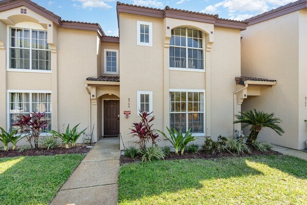 A two-story residential building with a beige exterior, numbered 872, featuring a front entrance with a door, windows, and a small garden with various plants. - 8702 Papaya DR, 2bed (Kissimmee)
